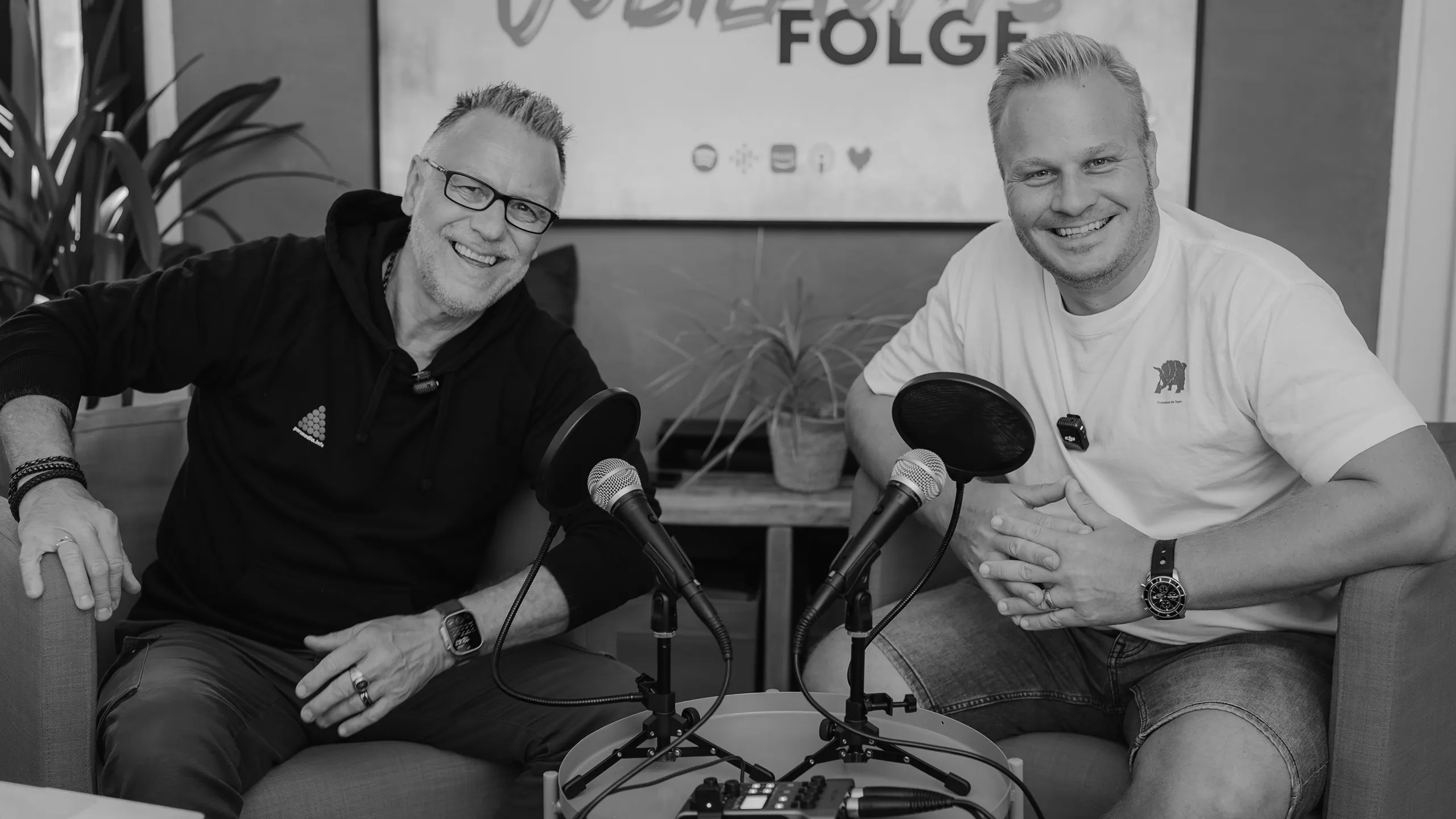 Zwei Podcaster sitzen im Studio vor Mikrofonen, lächelnd bei einer Podcast-Aufnahme, Hintergrundbanner „FOLGE“