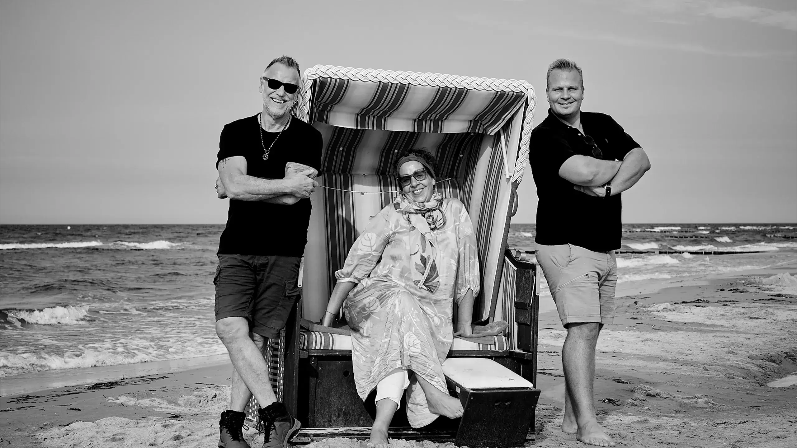 Geschäftsführung von parsmedia posiert vor dem Meer mit einem Strandkorb, im Hintergrund Wellen und Sandküste.Links vom Strandkorb steht Klaus Schenkmann, im Starndkorb sitzt Hilke Höfchen, Inhaberin von parsmedia und rechts steht Patrick Neumann.
