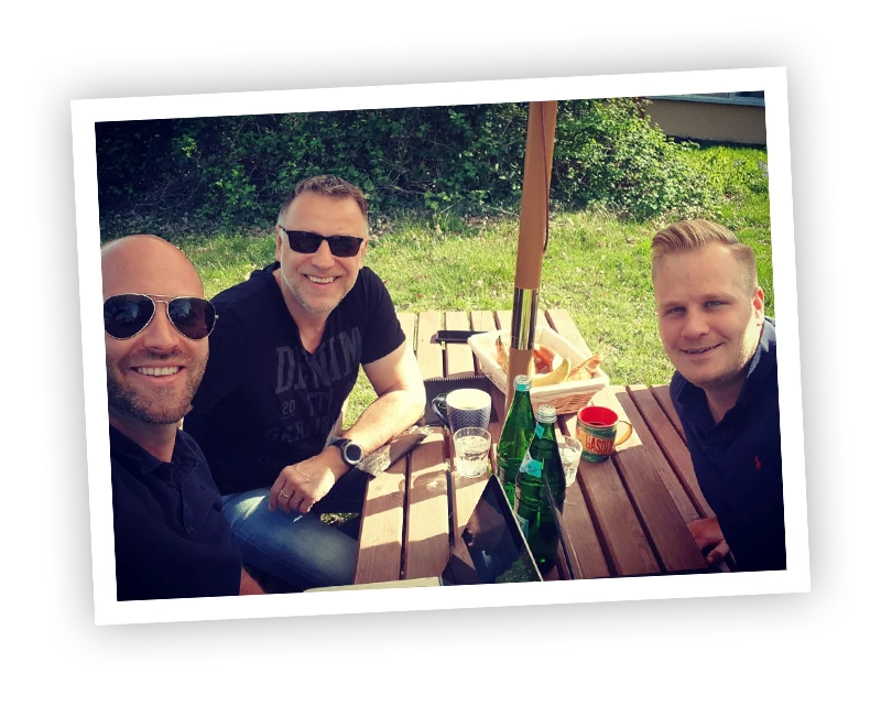 Drei Männer sitzen bei sonnigem Wetter am Picknicktisch, mit Bierflaschen und Snacks.